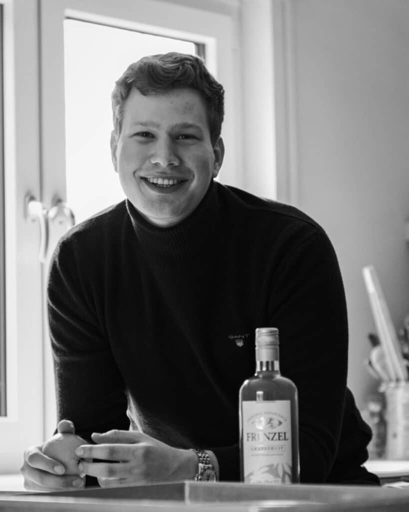 Johannes Frenzel, der Gründer, lächelt in die Kamera. Vor ihm steht eine Flasche FRENZEL Grapefruit, und er hält eine Grapefruit in den Händen.